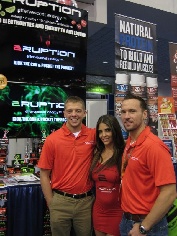 Two men and a woman in red Eruption shirts and dress pose together in front of Eruption effervescent energy displays and a Natural Protein banner.
