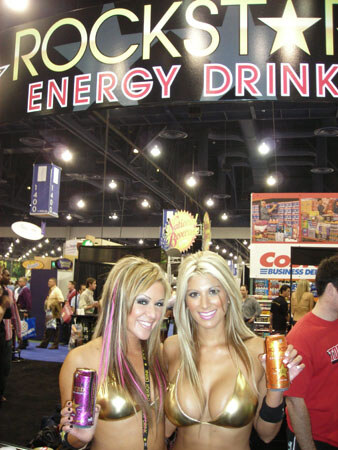 Two women in gold bikini tops hold Rockstar cans beneath a large Rockstar Energy Drink sign, with other exhibit booths and attendees visible behind them.