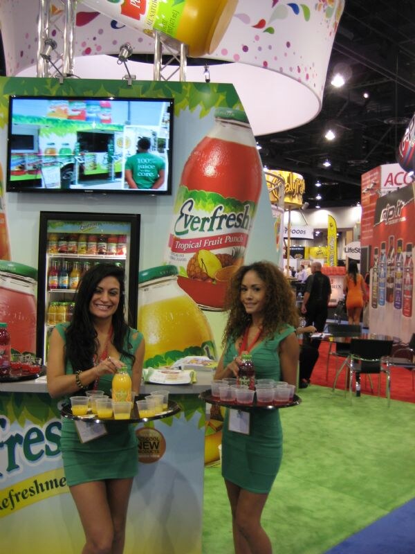 Two women in green dresses hold trays of juice samples in front of an Everfresh display with a large Tropical Fruit Punch bottle graphic and a refrigerated case.
