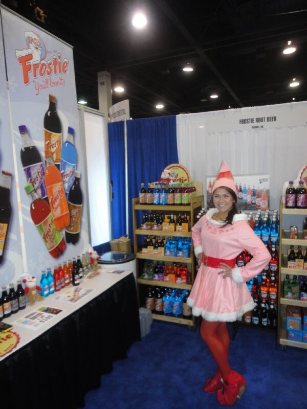 A woman in a pink elf costume with a pointed hat stands beside shelves of Frostie soda bottles and a Frostie banner at the Frostie Root Beer booth.