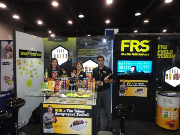 Two women and a man in dark shirts stand behind a counter of FRS drinks and chews. Signs advertise a chance to win a Tim Tebow autographed football.