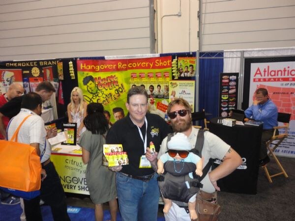 A man in a black shirt holds Hangover Recovery Shot packs beside a bearded man in sunglasses wearing a baby carrier. Atlantic Retail and Cherokee Brand signs appear behind them.