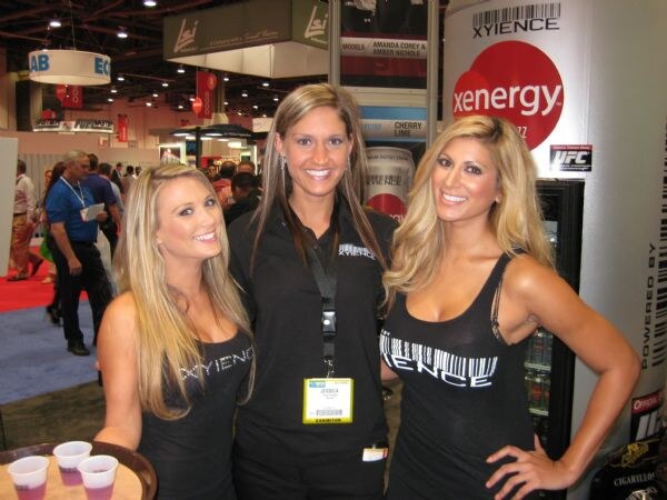 Three smiling women in black Xyience tops stand together; Jessica is in the center wearing an exhibitor badge. A Xenergy and Xyience display stands behind them, and one holds a tray of small cups.