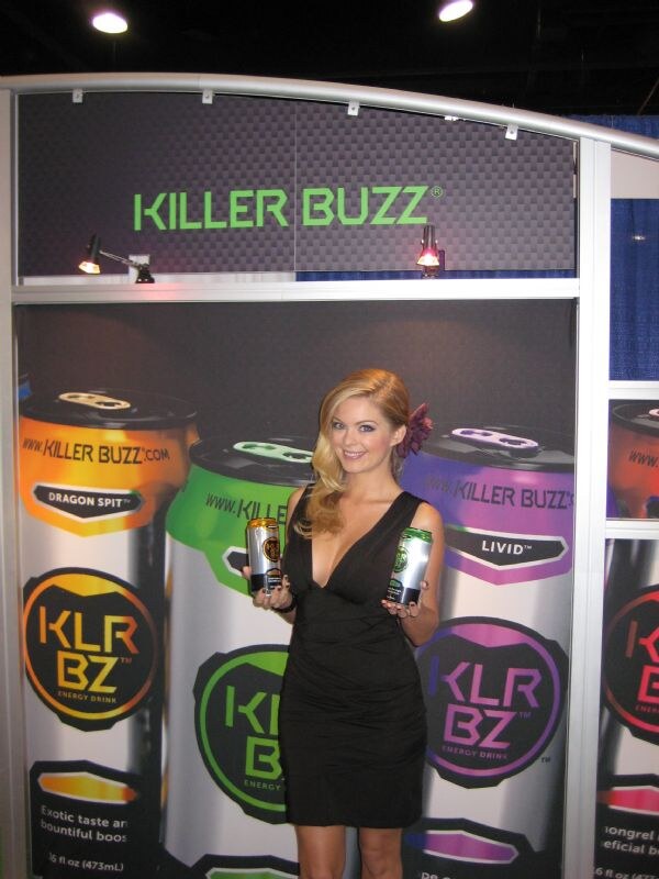 A blonde woman in a black dress holds two Killer Buzz cans in front of a KLR BZ backdrop showing large can graphics in several flavors.