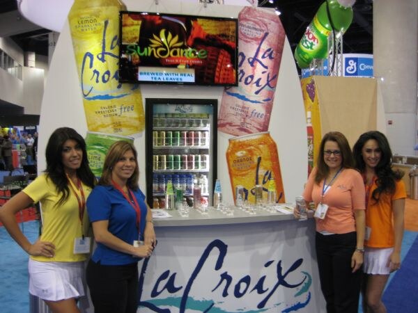 Four women stand around a LaCroix booth with large can graphics, a SunDance tea screen and a refrigerated case of drinks.