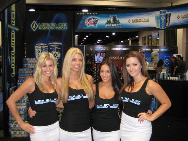 Four smiling women in black Liquid Ice Energy Drink tops and white skirts pose at the Liquid Ice Energy Drink booth, with stacked cases behind them and International Vapor Group and Smoke signs beyond.