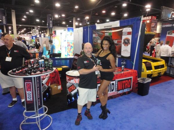 A bald man in a black polo and a woman in a black dress and boots stand in front of the Liquid Nitro booth, with branded displays, bottles on a round table and a yellow sports car nearby.