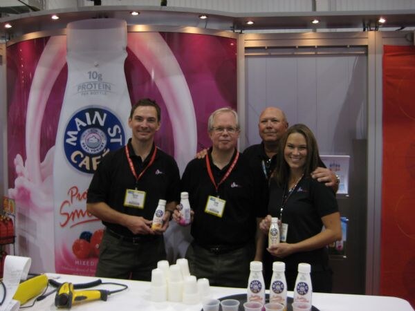 Three men and a woman in black shirts and lanyards stand behind a table at the Main St. Cafe booth, holding small bottles, in front of a large Main St Cafe protein smoothie bottle graphic.