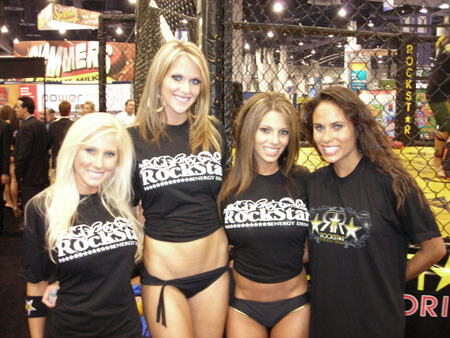 Four women in black Rockstar Energy Drink shirts stand together in front of a caged fighting ring display at a trade show.