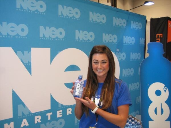 A smiling woman in a blue shirt holds a small Neo water bottle in front of a blue Neo Smarter Water backdrop, with a large blue Neo bottle display beside her.