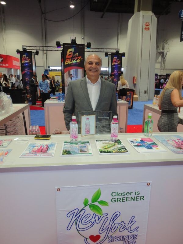 A smiling man in a gray blazer stands behind a counter with VBlast bottles and brochures, below a New York Springs USA sign, with 8 Hr Buzz banners behind him.