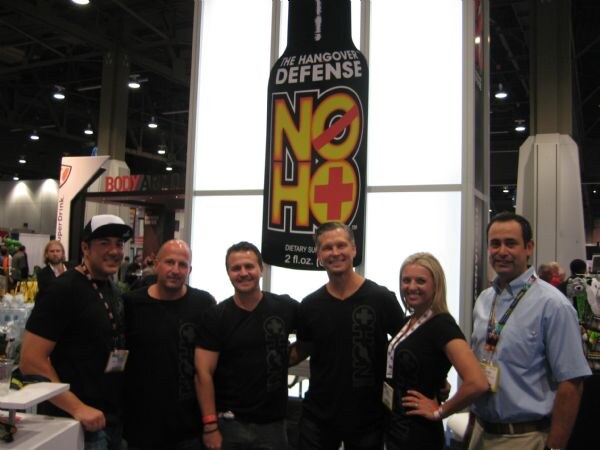 Five men and a woman, mostly in black shirts and wearing badges, stand together beneath a large NO HO The Hangover Defense bottle sign at the exhibit hall.