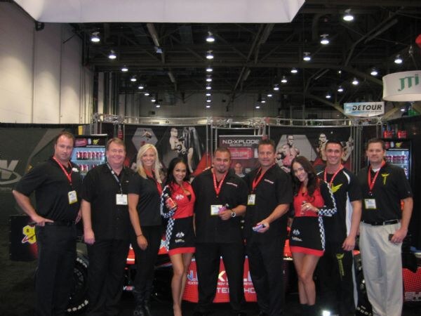 A group of men in black shirts and two women in red and checkered outfits pose in front of a race car and NO-XPLODE and Detour signs inside a convention hall.