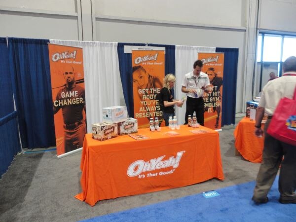 A man and a woman stand behind an orange table with an Oh Yeah! tablecloth, with product boxes and bottles on top and orange Oh Yeah! banners behind them. A visitor stands at right.