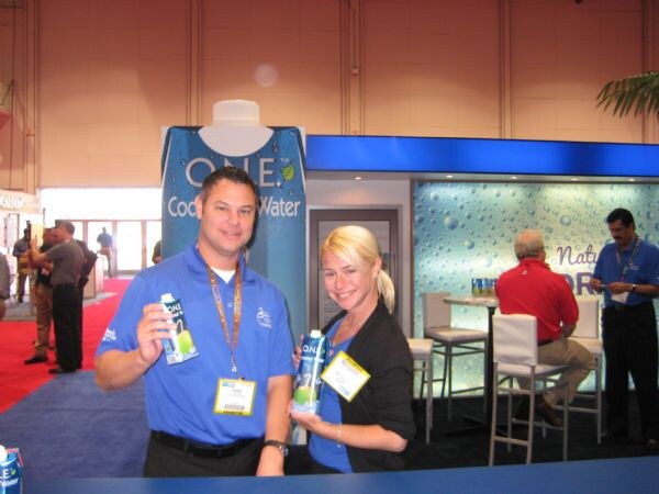 A man in a blue polo and a blonde woman each hold a carton of O.N.E. Coconut Water in front of a tall O.N.E. Coconut Water display, with other attendees in the background.