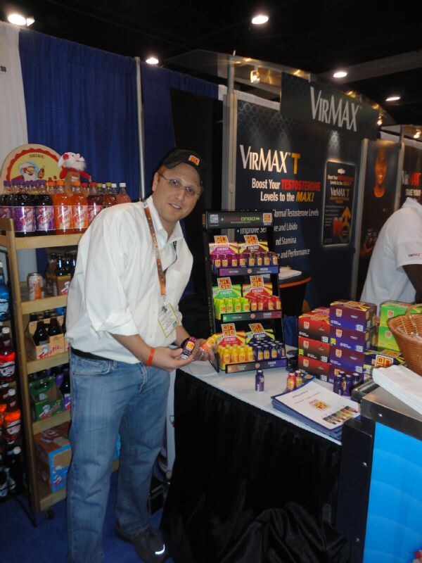 A man in a white shirt, glasses and cap holds a small bottle beside an Ongo shot display, with a VirMax T sign behind him and boxes of product on the table.