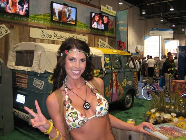 A smiling woman with a flower headband and peace-sign bikini top flashes a peace sign in front of a painted vintage van and Peace Tea signage with video screens.