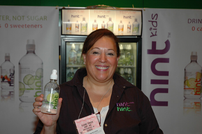 Elaine Triber of Hint, Inc. smiles while holding a bottle of Hint water in front of a lit refrigerator and Hint and Hint Kids banners.