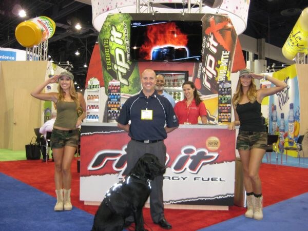 Two women in camouflage shorts salute beside a Rip It Energy Fuel counter where a man in a navy polo, two other people and a black dog stand.