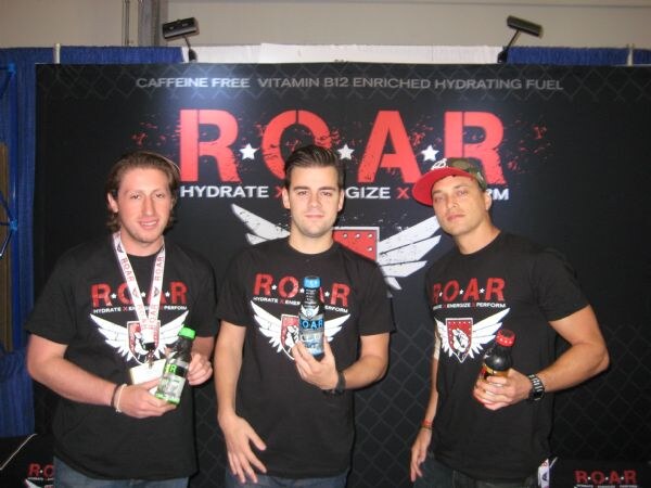Three men in black R.O.A.R. T-shirts hold bottles in front of a black R.O.A.R. banner reading Hydrate, Energize, Perform.