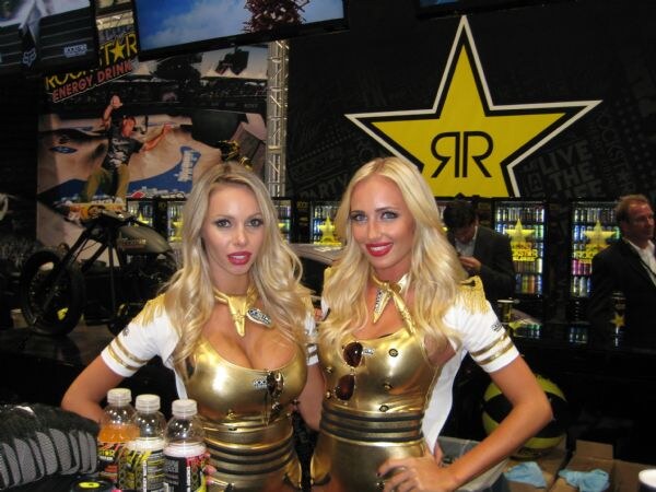 Two blonde women in gold outfits stand at the Rockstar booth, with a large yellow star logo, a motorcycle and lit drink coolers behind them.