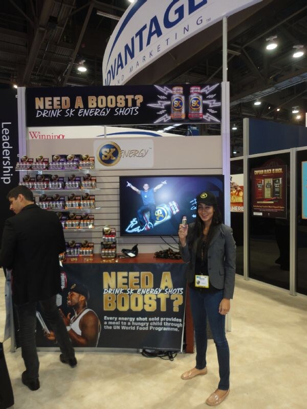 A woman in a black cap and gray blazer holds up an SK Energy shot beside a display reading Need a Boost? Drink SK Energy Shots; a man in black stands at left.