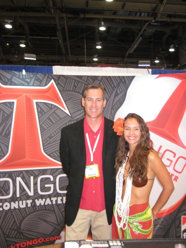 A smiling man in a black blazer and red shirt with a badge stands beside a woman with a flower in her hair and shell necklace, in front of a Tongo Coconut Water banner.