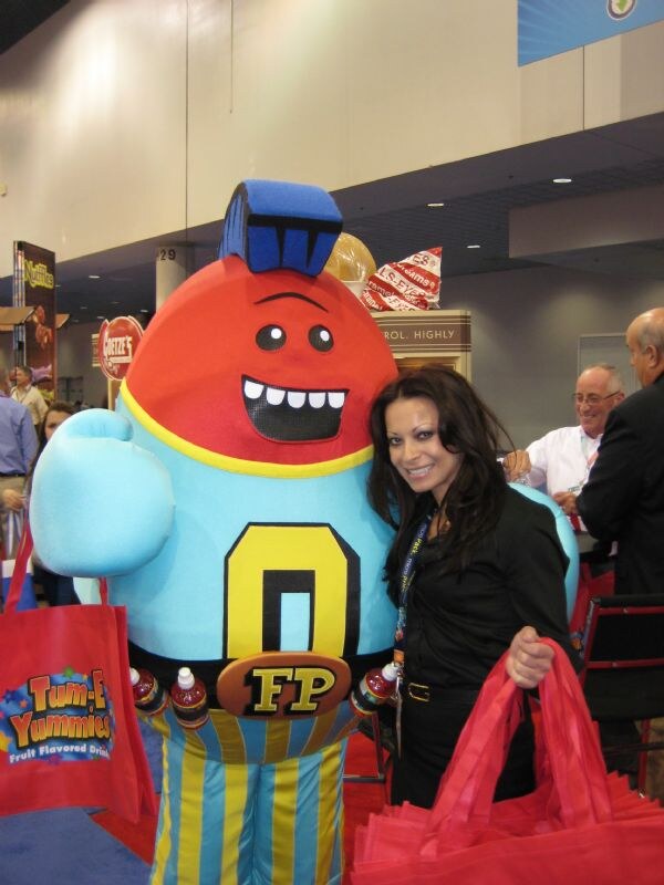 A woman in black holding red bags stands beside a large costumed mascot with a red head and blue body; a Tum-E Yummies Fruit Flavored Drinks bag is at left.
