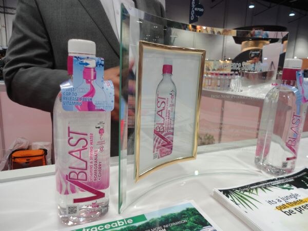 A VBlast pomegranate and cherry vitamin water bottle stands beside a framed glass award showing a VBlast bottle on a counter, with a person in a gray jacket behind.