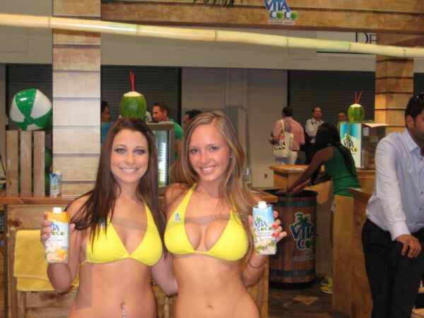 Two smiling women in yellow bikini tops hold Vita Coco coconut water cartons at a wooden booth with a beach ball and coconuts with straws.