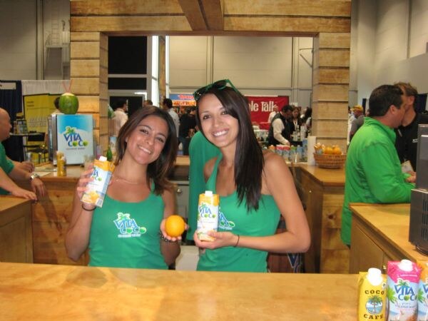 Two smiling women in green Vita Coco tank tops hold cartons of coconut water, one also holding an orange, behind a wooden counter.