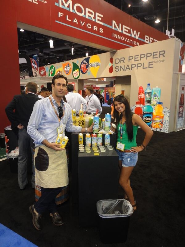 A man in a striped shirt and apron holds Vita Coco cartons beside a woman in a green top at a display table, with the Dr Pepper Snapple Group booth and bottles behind.