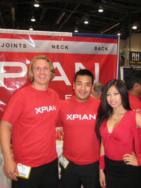 Two men and a woman in red Xpian shirts pose together in front of a red Xpian banner reading Joints, Neck, Back. An RH Labs sign is visible at right.