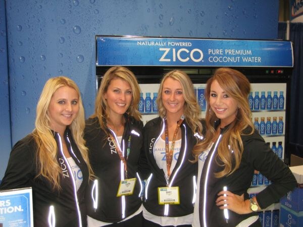 Four smiling women in black ZICO jackets stand in front of a ZICO Pure Premium Coconut Water cooler. Mary, second from right, wears a badge on a lanyard.