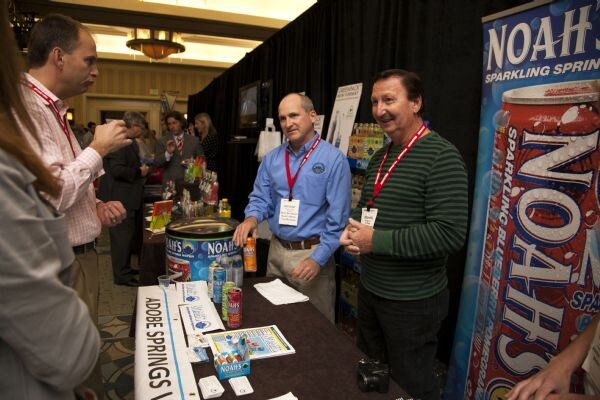 Two men in lanyards stand at a table of Noah's Sparkling Springs cans, talking with a man in a pink shirt at left. A tall Noah's banner stands at right and other attendees are in the background.