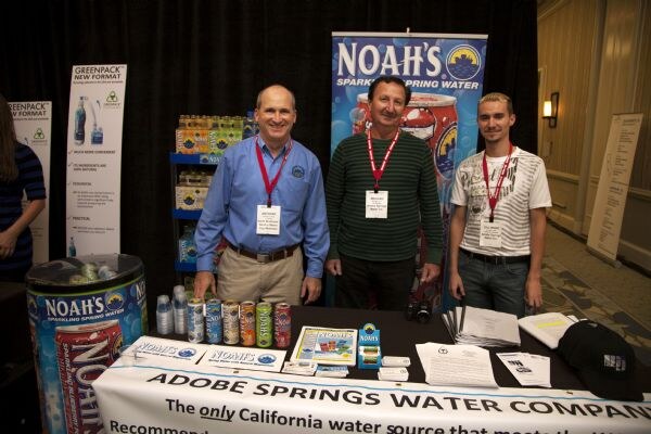 Three men with red lanyards stand behind a table with Noah's Sparkling Spring Water cans and a banner reading Adobe Springs Water Company. A Noah's banner and product shelf are behind them.