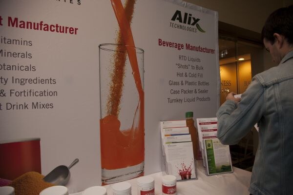An Alix Technologies display lists beverage manufacturing services over a table of brochures and sample containers. A person in a denim jacket stands at right, seen from the side.