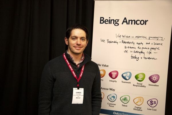 A smiling man with dark hair in a dark sweater and red lanyard stands in front of a white Being Amcor banner listing company values.
