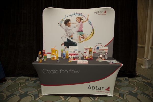 Aptar booth with a curved graphic backdrop of two children jumping, and a table displaying beverage bottles and brochures. The table cover reads "Create the flow" and Aptar food + beverage.
