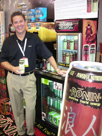 A smiling man in a black shirt and lanyard holds a Ronin can beside a Ronin refrigerated cooler and an oversized Ronin can display.