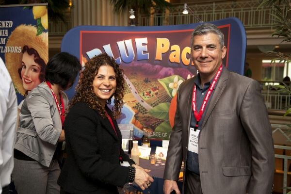 A smiling curly-haired woman in a black jacket and a smiling man in a gray blazer, both wearing red lanyards, pose at the Blue Pacific Flavors booth. Another woman stands behind them.