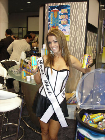 A smiling woman in a white corset top and black skirt wears a Royal Blunts sash and holds a can. A tub of cans sits to the right and people talk at a table behind.