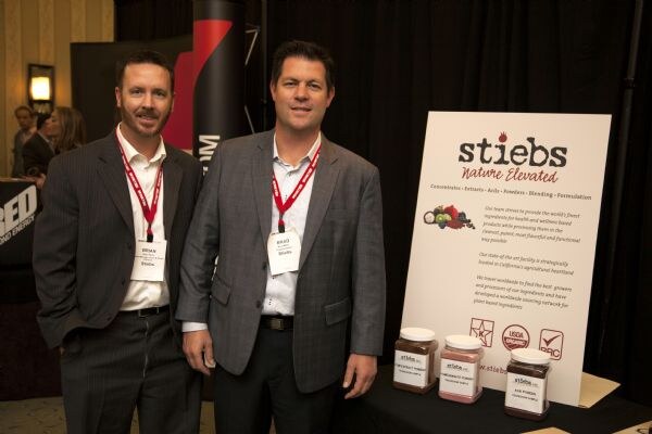 Brian and Brad of Stiebs, wearing suit jackets and red lanyards, stand smiling beside a stiebs Nature Elevated sign and jars on a table.