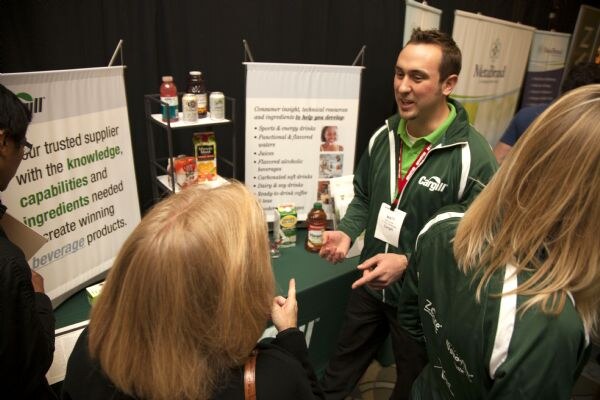 A man in a green Cargill jacket gestures while talking to two attendees in the foreground at the Cargill booth. Banners and a shelf of beverage samples stand behind him, with a NetaBrand banner nearby.