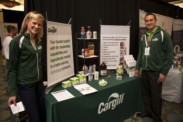 A woman and a man in green Cargill jackets stand beside a green-covered Cargill table with beverage samples, brochures and banners about ingredients for beverage development.