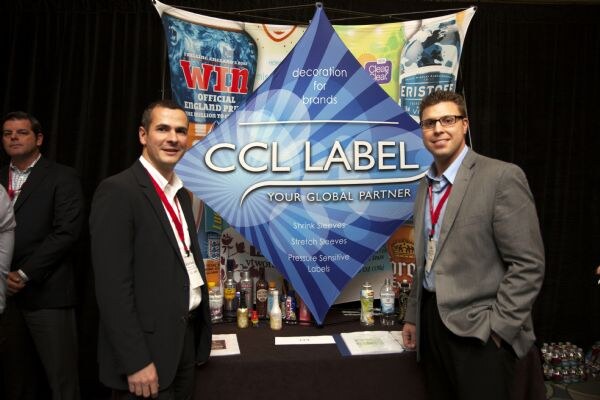 Two men in blazers wearing red lanyards stand in front of a CCL Label booth. The sign reads "Your global partner" and lists shrink sleeves, stretch sleeves and pressure sensitive labels. Bottles sit on the table.