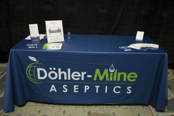 A blue table cover printed with the Döhler-Milne Aseptics logo, with brochures, pens and a small sign on top, in front of a black curtain.
