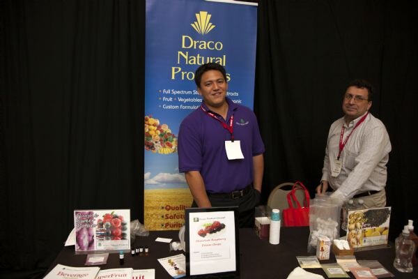 Two men wearing red lanyards stand behind a table at the Draco Natural Products booth, in front of a blue banner. The table holds product literature, small bottles and sample items.