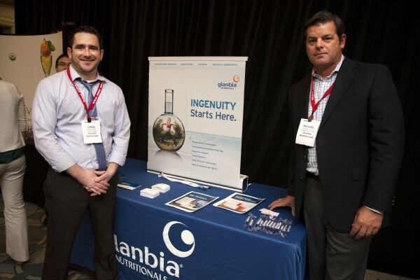 Two men wearing red lanyards and name badges stand beside a blue-covered Glanbia Nutritionals table. A banner reads "Ingenuity Starts Here." Brochures and cards lie on the table.