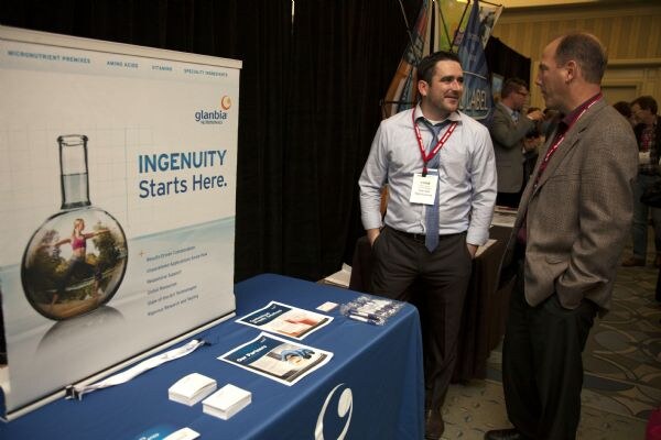 A man in a blue shirt and tie talks with a man in a tweed jacket beside the Glanbia Nutritionals booth, which has an "Ingenuity Starts Here." banner and a table with brochures.